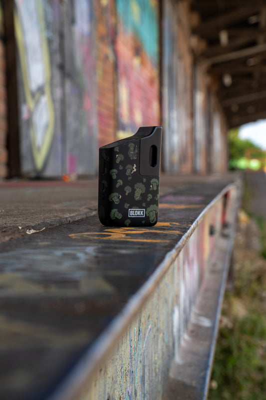 Schwarzer BLOKK Vaporizer mit Brokkoli-Design steht auf einer bemalten Betonfläche vor einer urbanen Graffiti-Kulisse.